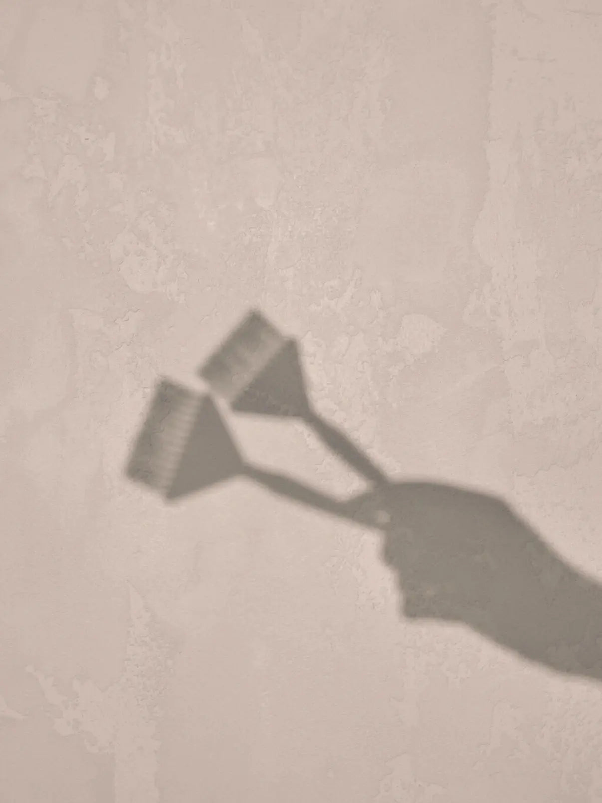 Artistic shadow silhouette of a hand holding two Foxy Blondes hair colouring brushes, cast onto a textured light beige-pink wall, creating a minimalist design.
