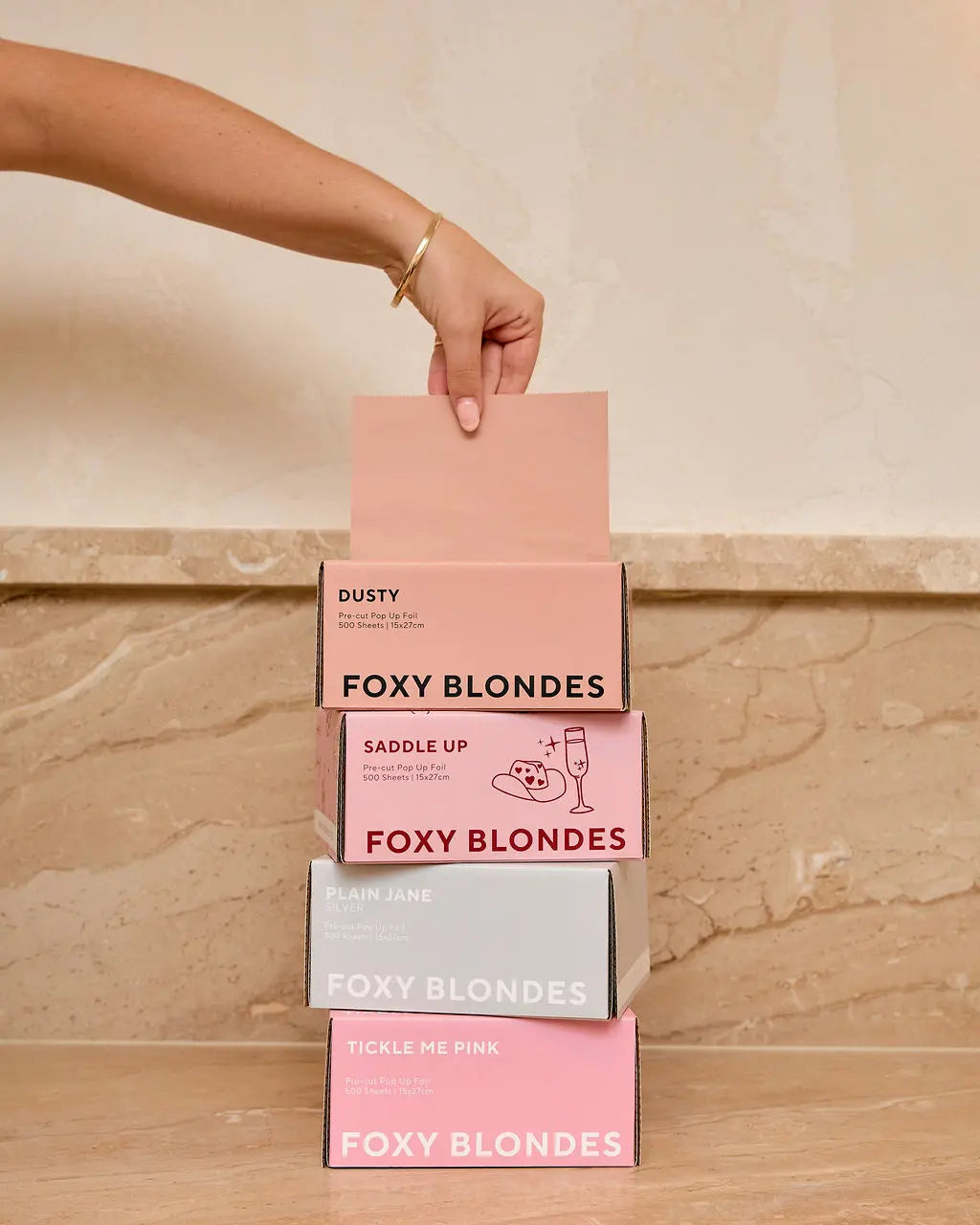 Hand dispensing a foil from a stack of four Foxy Blondes pre-cut pop-up hair foil boxes: 'Dusty' (dusty rose), 'Saddle Up' (pink patterned), 'Plain Jane Silver', and 'Tickle Me Pink', showcasing the product range.