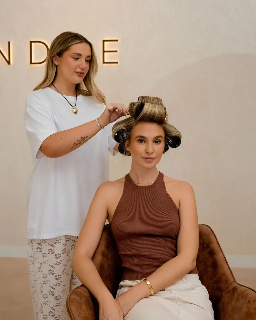 Hairstylist setting large black Foxy Blondes hair rollers in a blonde client's hair for a voluminous style in a modern salon.
