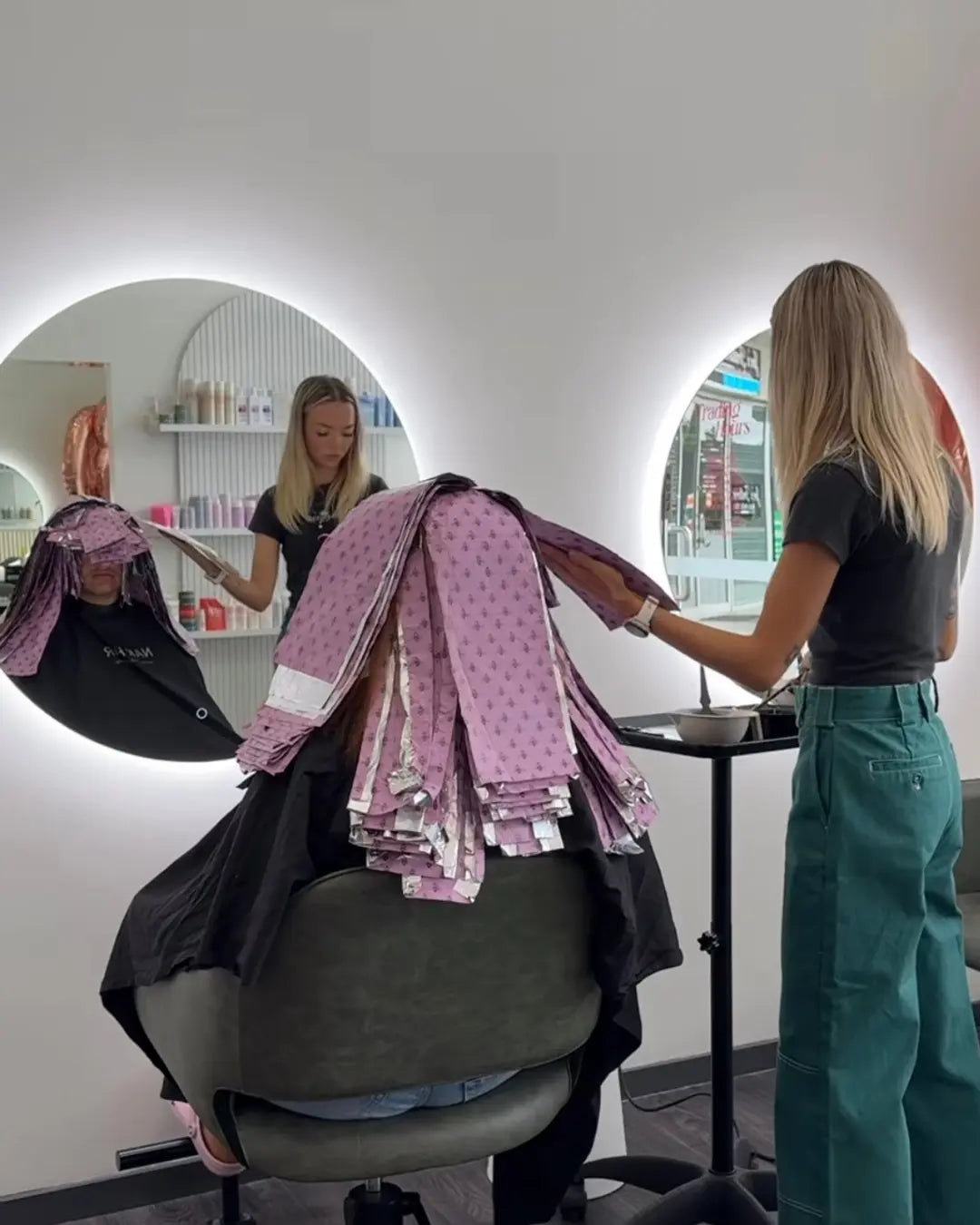 Hairstylist applying Foxy Blondes oh honey patterned hair foils for a full head highlighting service on a client in a modern salon with large round backlit mirrors.