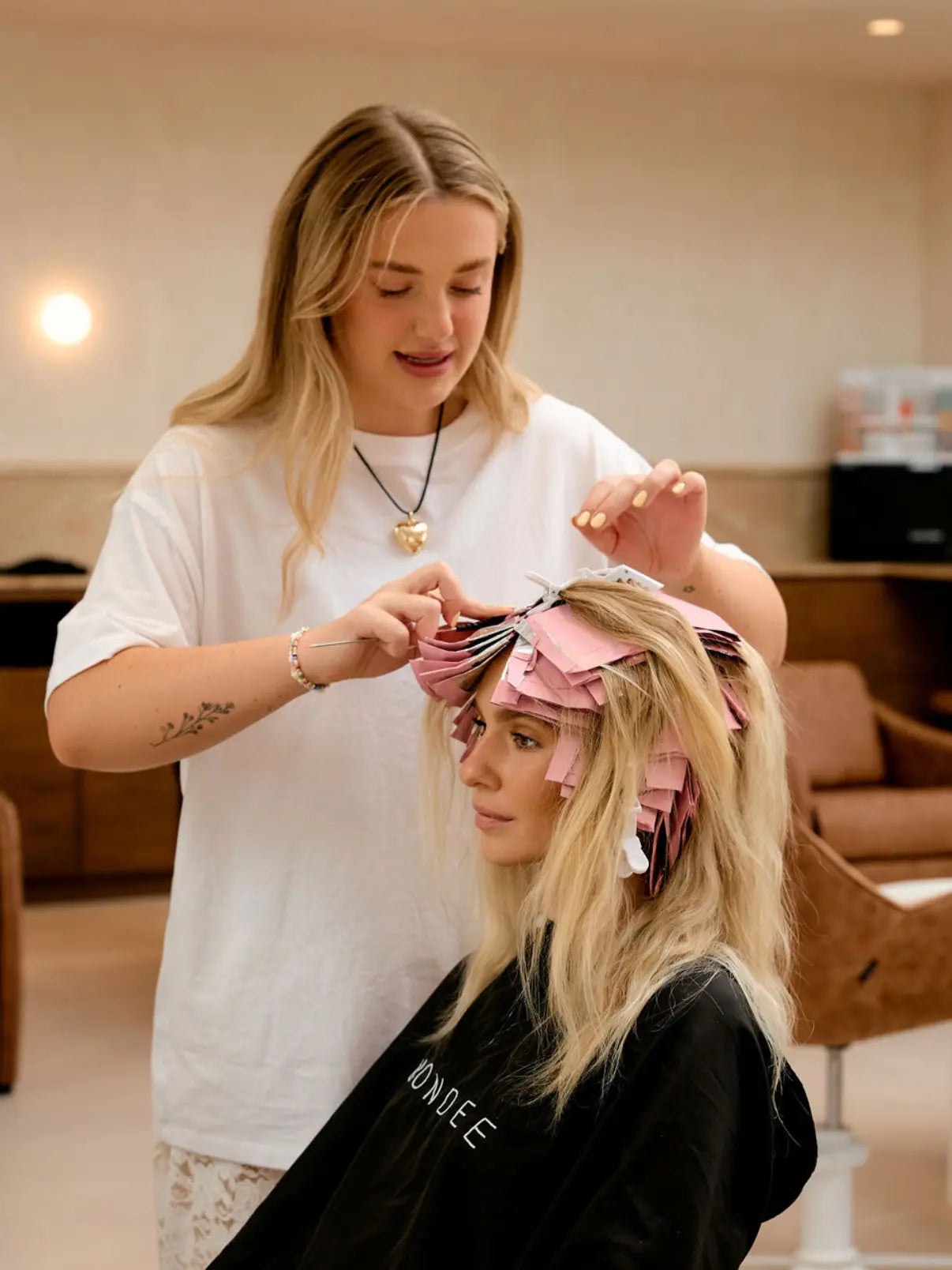 Professional hairstylist meticulously applying Foxy Blondes pink hair foils to a blonde client wearing a salon cape during a highlighting service.