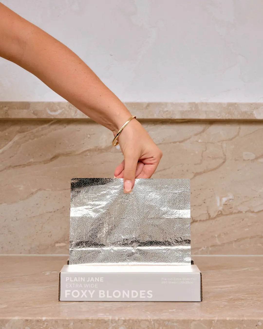 Hand pulling a Foxy Blondes 'Plain Jane' Extra Wide silver embossed pre-cut hair foil (20x35cm) from its 200-sheet dispenser box on a marble surface.