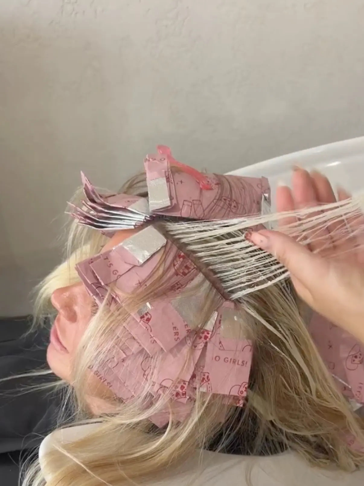 Close-up of Foxy Blondes 'Saddle Up' pink patterned hair foils (Let's Go Girls! theme) applied to blonde hair, with a stylist combing through a finely highlighted section to check processing at a salon washbasin.