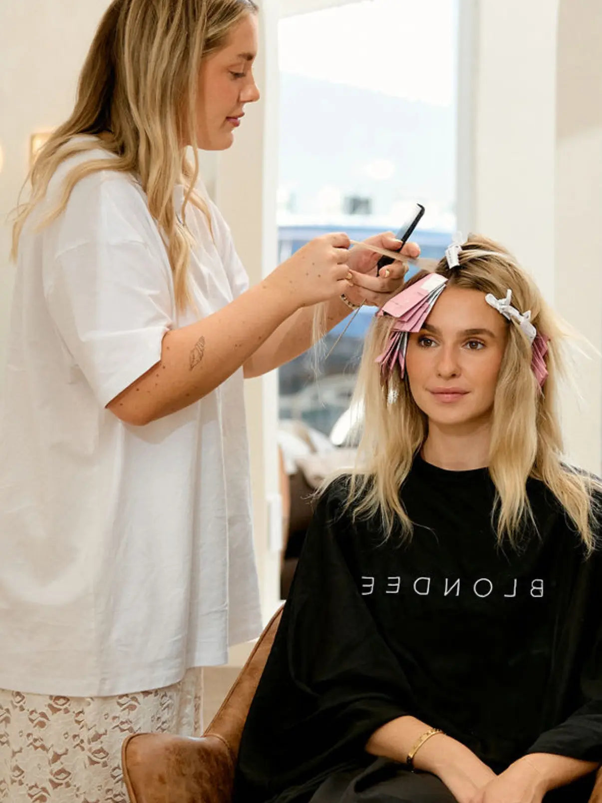 Professional hairstylist applying Foxy Blondes 'Tickle Me Pink' hair foils to a blonde client's hair during a highlighting service in a bright salon.