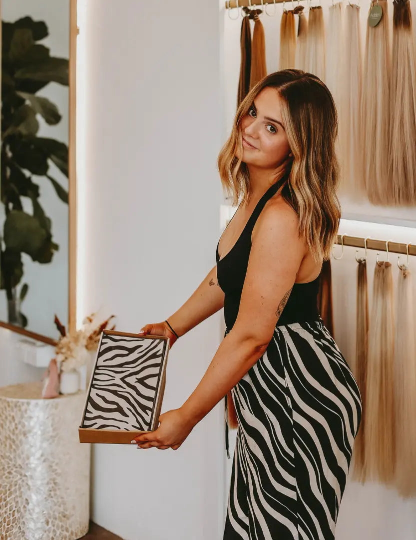 Stylish hairstylist holding an open box of Foxy Blondes zebra print hair foils in a modern salon, with hair extensions displayed in the background and wearing a matching zebra print skirt.