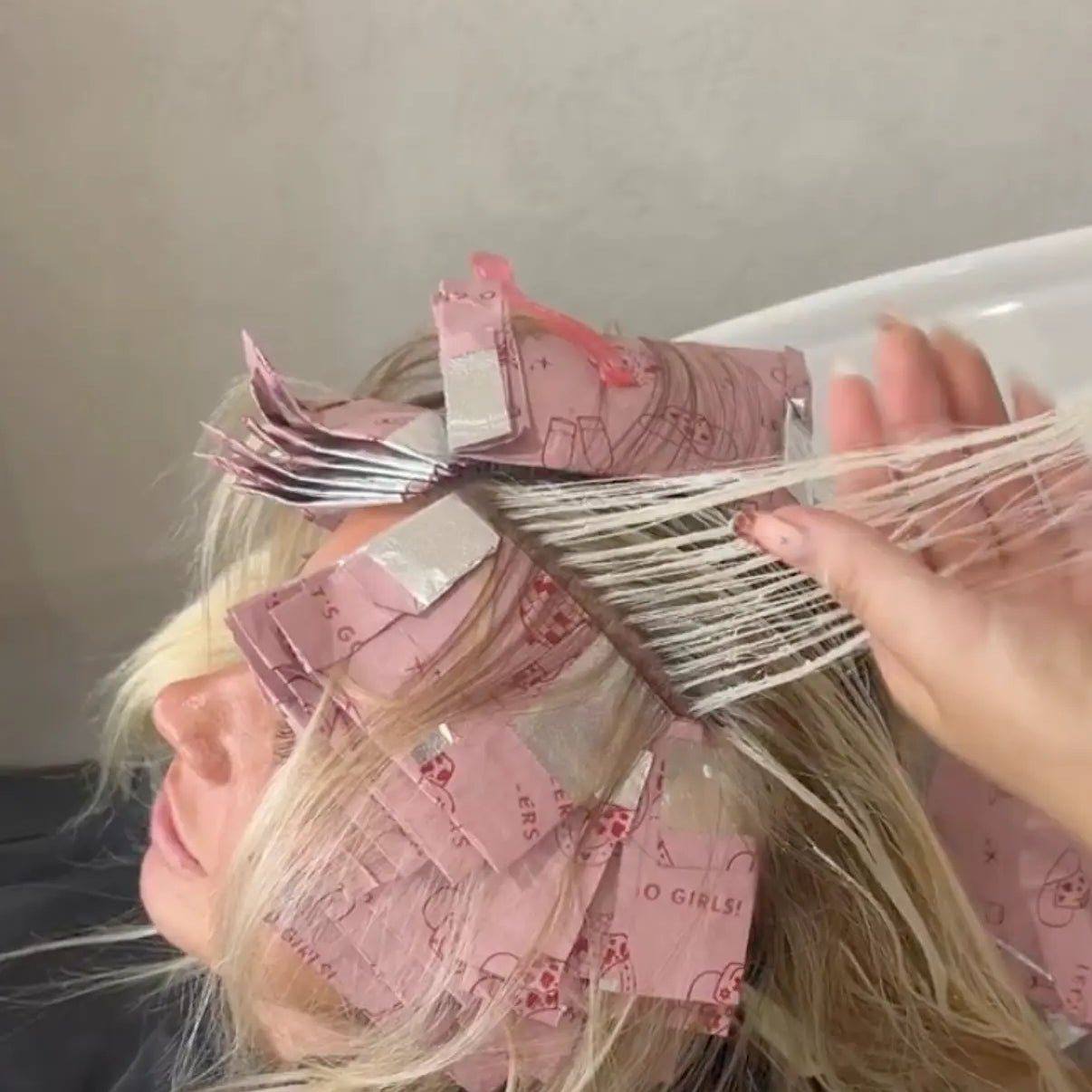 Close-up of Foxy Blondes 'Saddle Up' pink patterned hair foils (Let's Go Girls! theme) applied to blonde hair, with a stylist combing through a finely highlighted section to check processing at a salon washbasin.